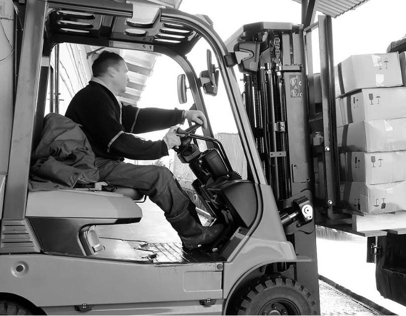 Een magazijnmedewerker bedient een vorkheftruck en tilt een pallet met dozen omhoog, in een industriële omgeving.